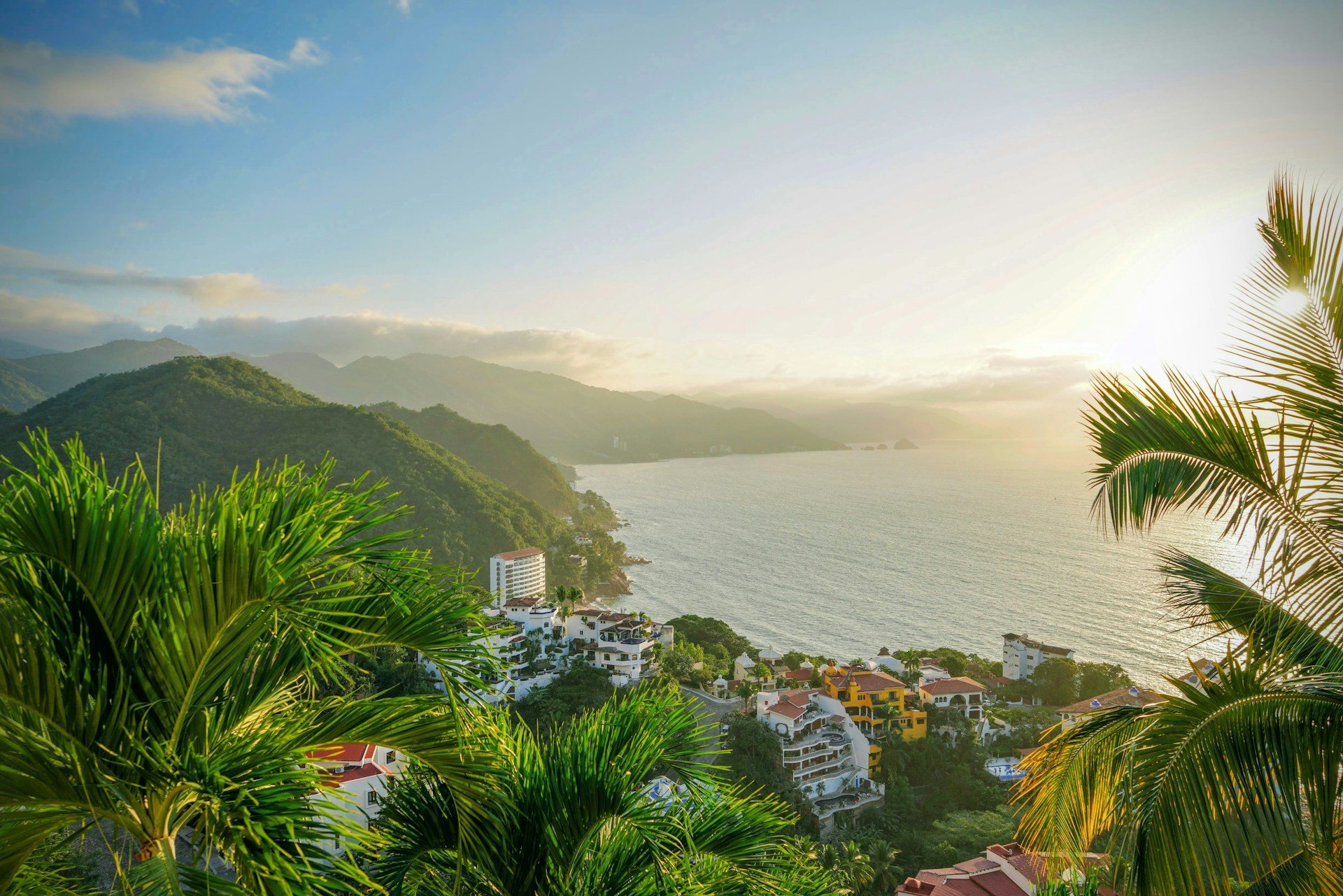 Blick zwischen Palmen auf eine weite Bucht mit grünen Bergen und weißen Häusern am Hang (Symbolbild, nicht Bali)