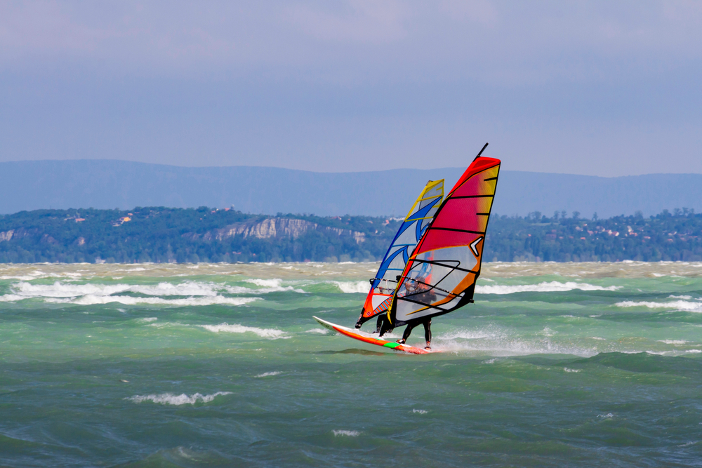 Δύο windsurfer με σανίδες χωρίς foil τρέχουν με δυνατό αέρα πάνω σε αφρισμένο νερό λίμνης, με δάσος και άσπρο βράχο στην ακτή· ενδεικτική εικόνα, δεν είναι από την Κρήτη