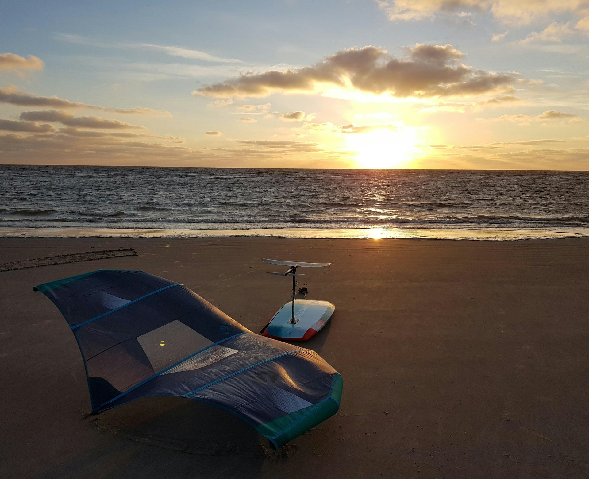 Una wing tendida sobre la arena y, detrás, una tabla con el foil hacia arriba, frente al mar al atardecer (imagen ilustrativa)