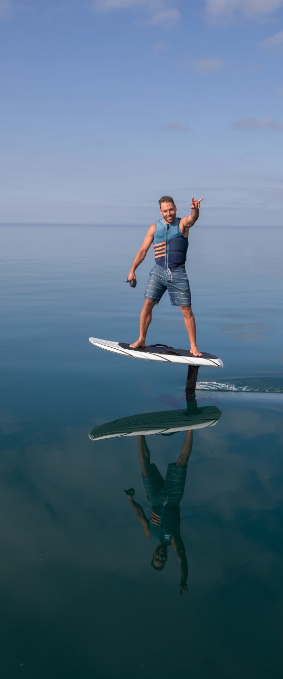 Rider sur un eFoil en vol, télécommande en main, main levée, reflet sur une eau plate (photo d’illustration, pas prise en Vendée)