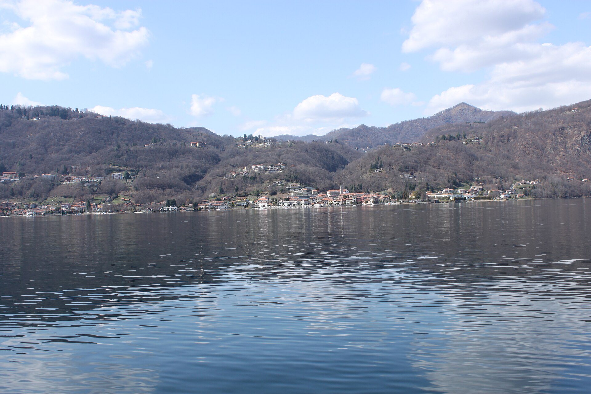 Le lac d’Orta par temps clair, eau calme au premier plan et village aux toits rouges au pied de collines boisées encore sans feuilles