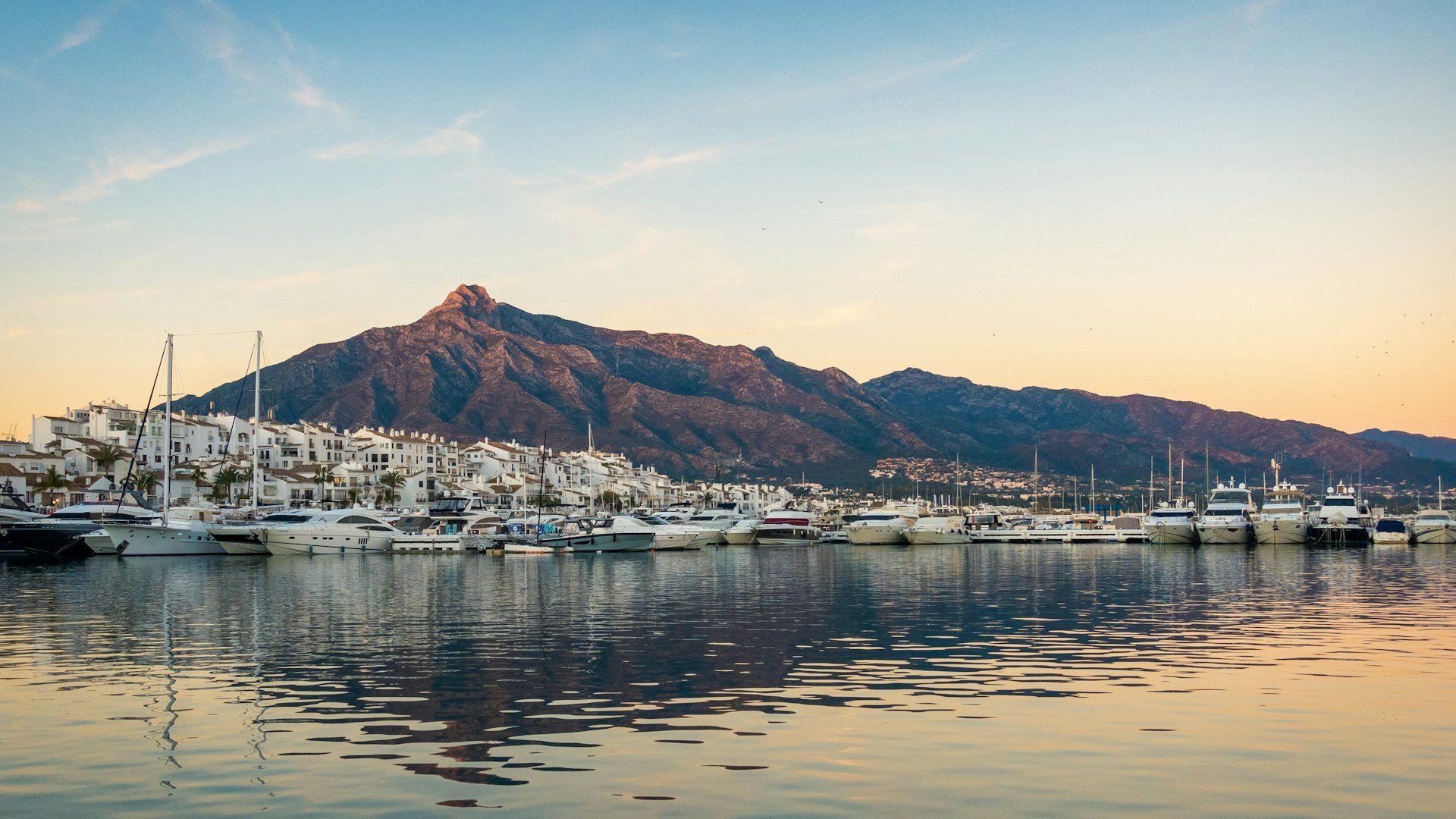 La marina de Puerto Banús à Marbella au coucher du soleil, avec des yachts à quai, des immeubles blancs et la montagne de La Concha derrière