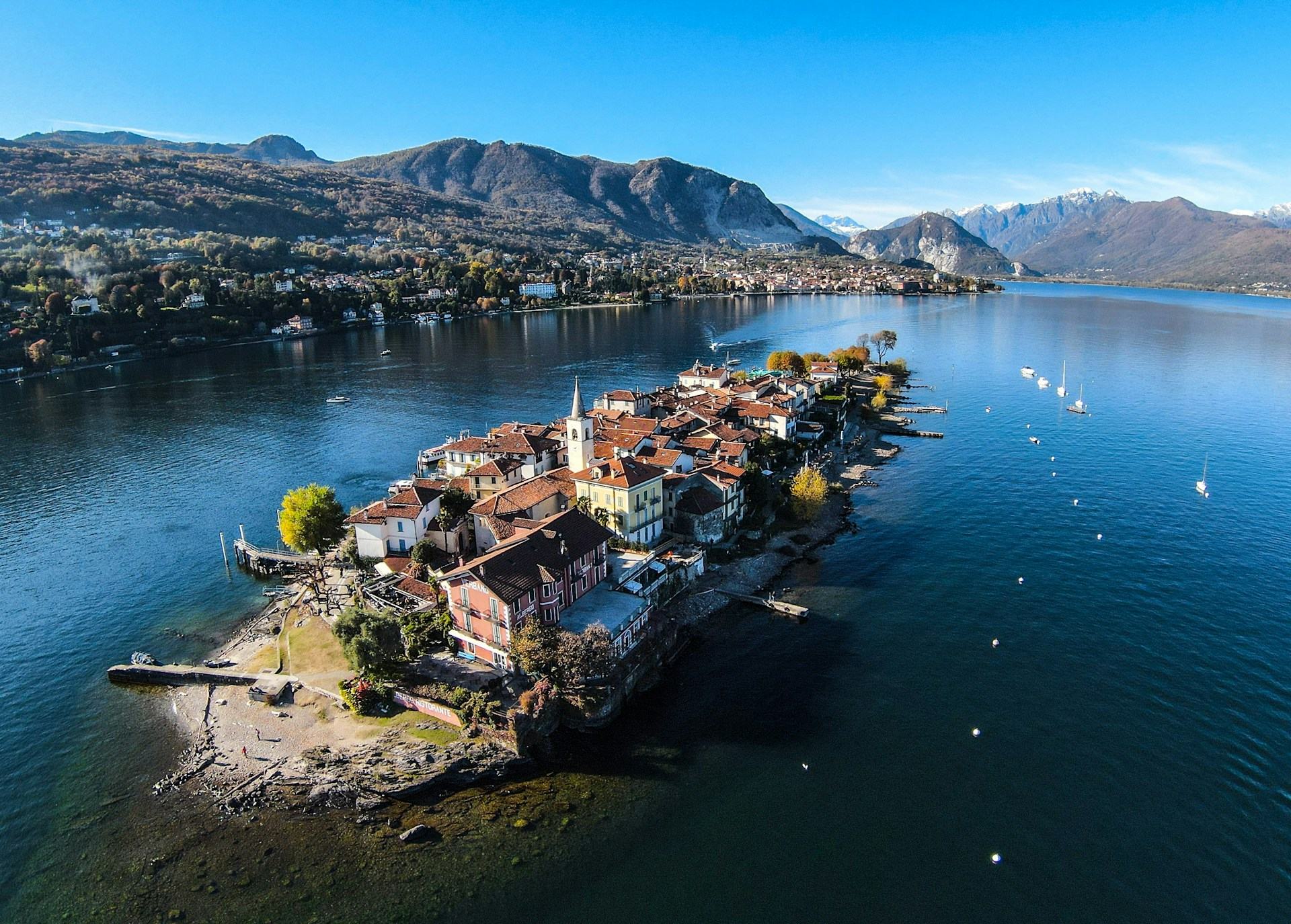 L’Isola dei Pescatori vista dall’alto, con le case dai tetti rossi, il campanile e i pontili, e dietro la riva e le montagne intorno al lago
