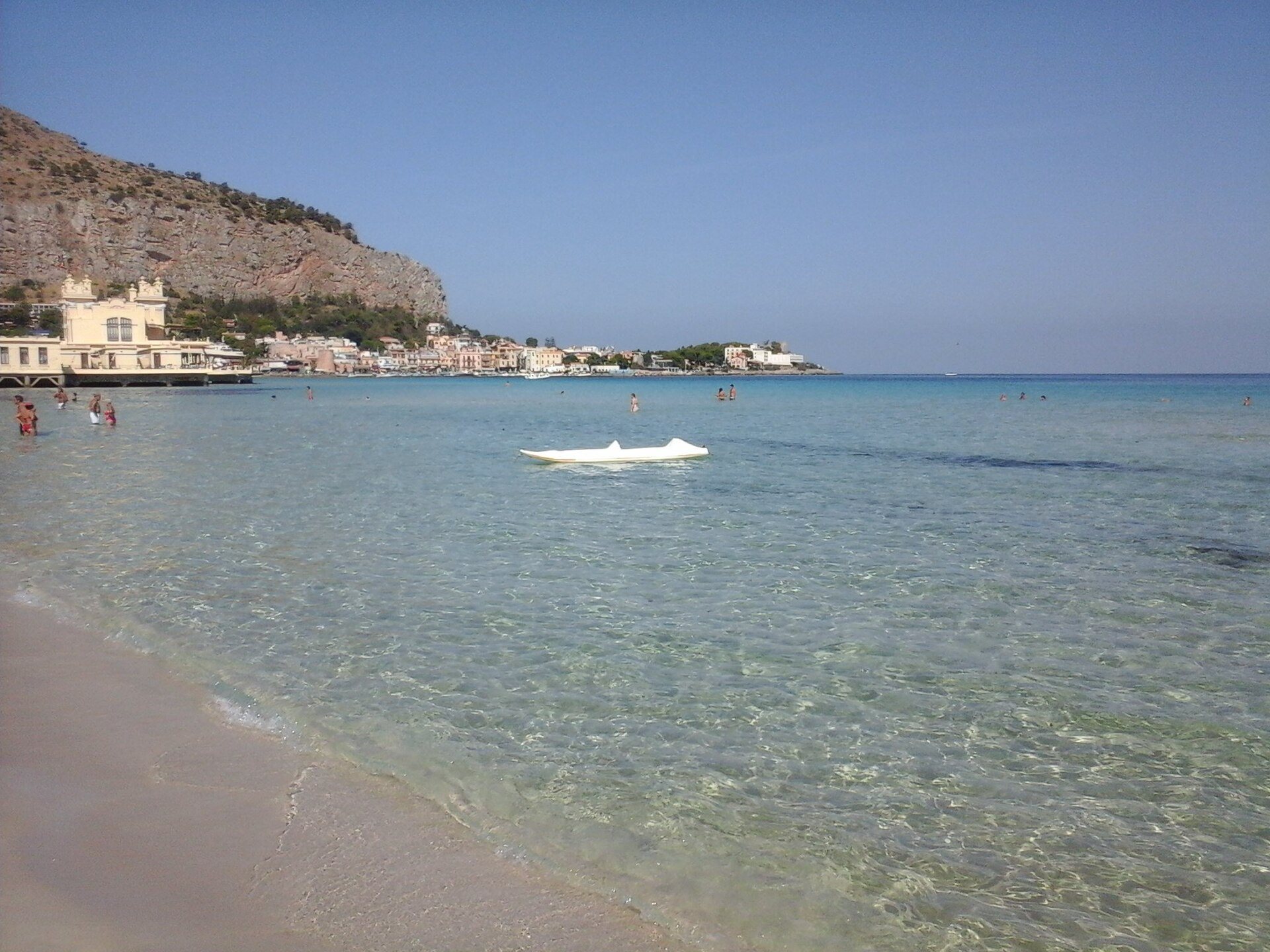 La spiaggia di Mondello a Palermo, con l’acqua bassa e trasparente sulla sabbia, bagnanti in mare, una piccola barca bianca e lo stabilimento liberty sotto la montagna
