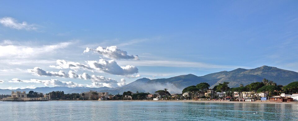 Panorama del golfo di Mondello con l’acqua calma in primo piano, un edificio liberty sul mare a sinistra, la riva con pini e case e le montagne sotto un cielo di nuvole