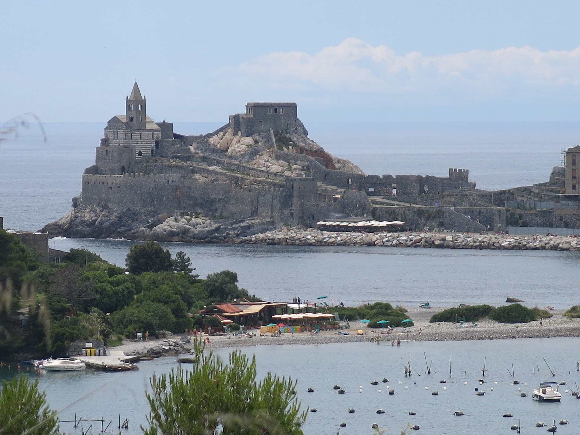 Spiaggia di ciottoli con ombrelloni e barche all’ormeggio sull’isola Palmaria, con Porto Venere e la sua chiesa sul promontorio al di là dello stretto