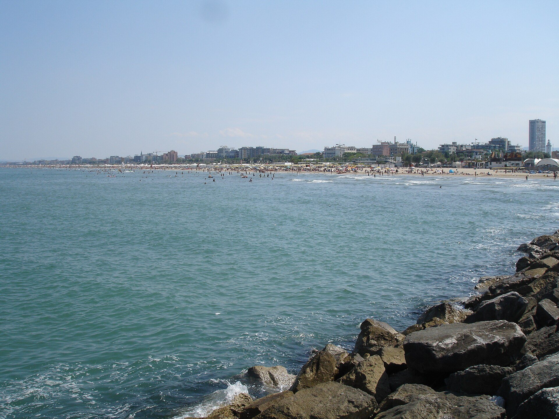 La spiaggia di Rimini vista dagli scogli di un frangiflutti, con bagnanti in acqua, file di ombrelloni e gli edifici del lungomare