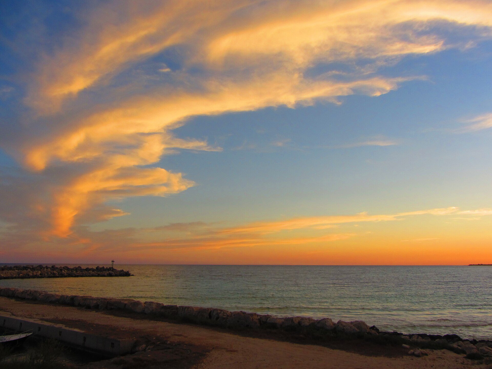 Tramonto sul mare a Santa Maria di Leuca, con nuvole arancioni nel cielo, un molo di scogli a sinistra e la riva di pietre in primo piano