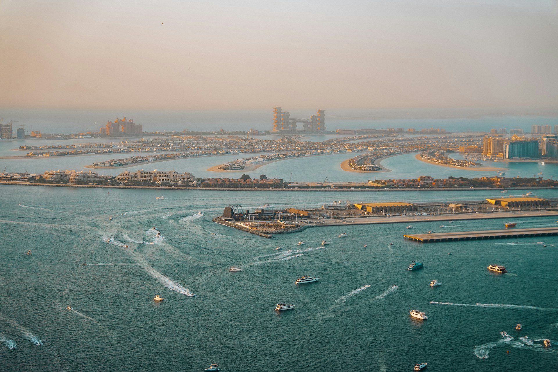 Luchtfoto van de kust van Dubai in avondlicht, met de buitenste ring en de bladeren van Palm Jumeirah aan de horizon en op de voorgrond zee met veel boten en jachten