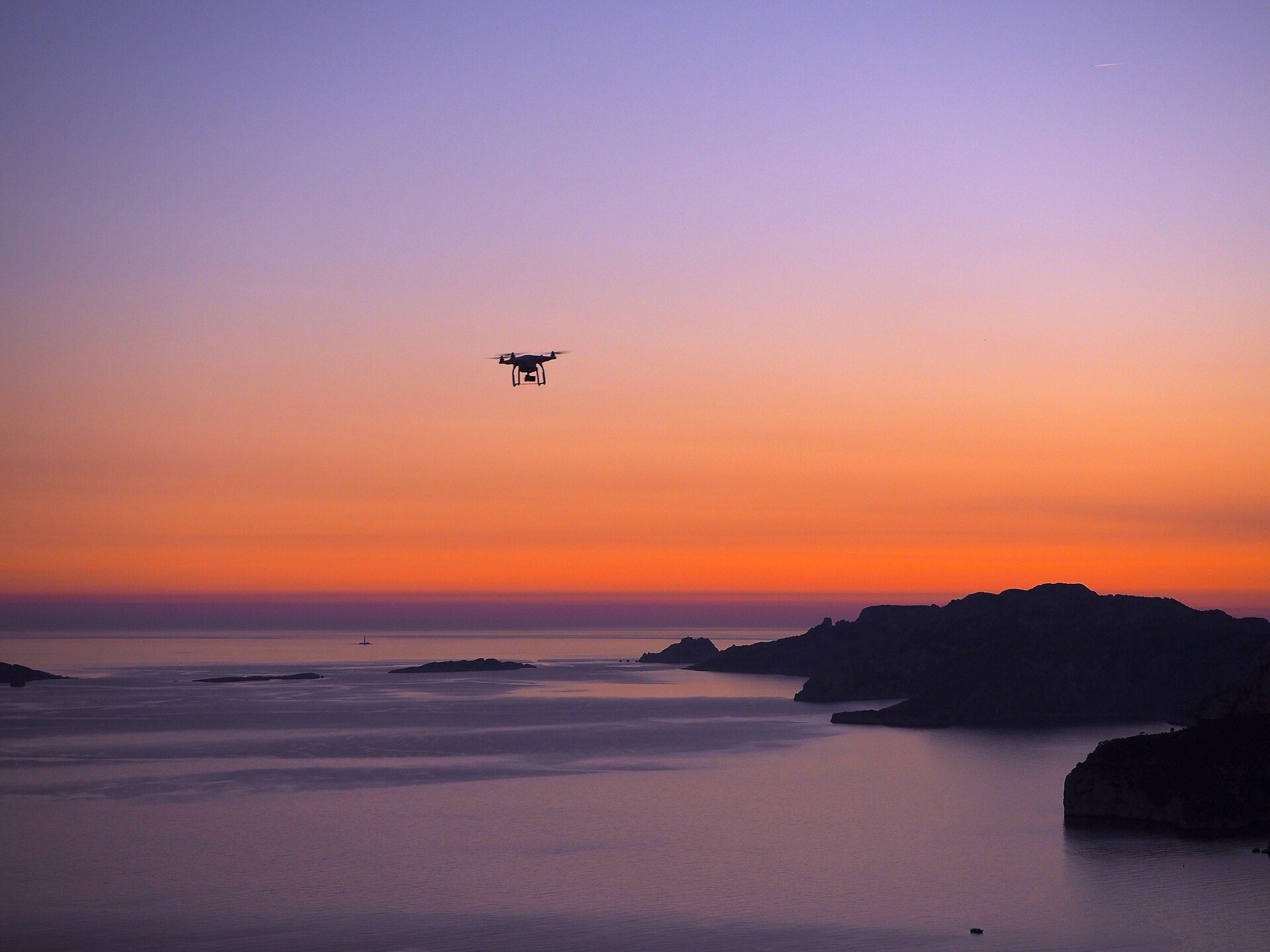 Een drone boven een kalme zee bij zonsondergang, met een rotskust en eilandjes in silhouet (beeld ter illustratie, geen foto van de Bretonse kust of van een school op deze pagina)