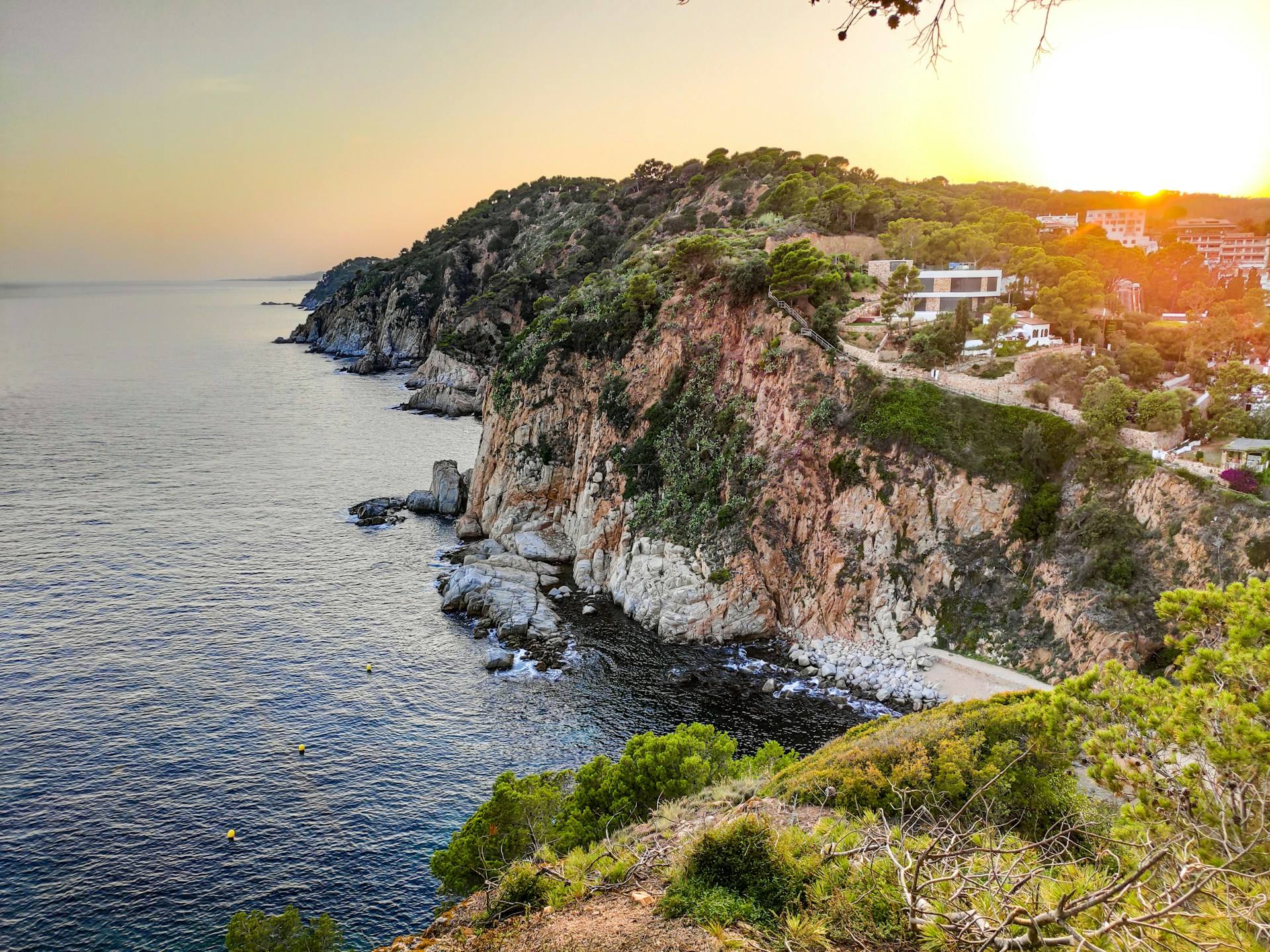 Rotskust met pijnbomen bij Tossa de Mar aan de Costa Brava bij zonsondergang, met villa’s op de helling en gele boeien in het water