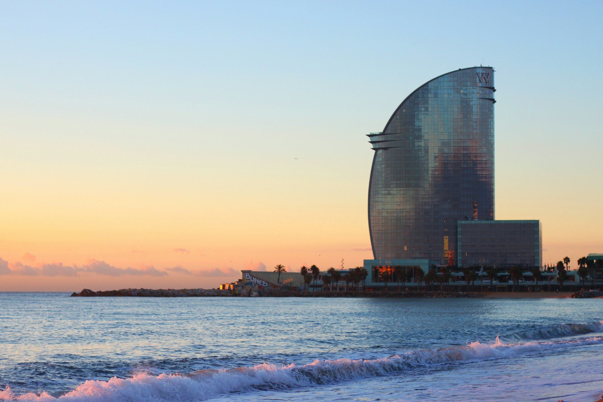 Plaża Barceloneta o wschodzie słońca, w tle szklany hotel w kształcie żagla przy wejściu do portu; na wodzie nie ma deski