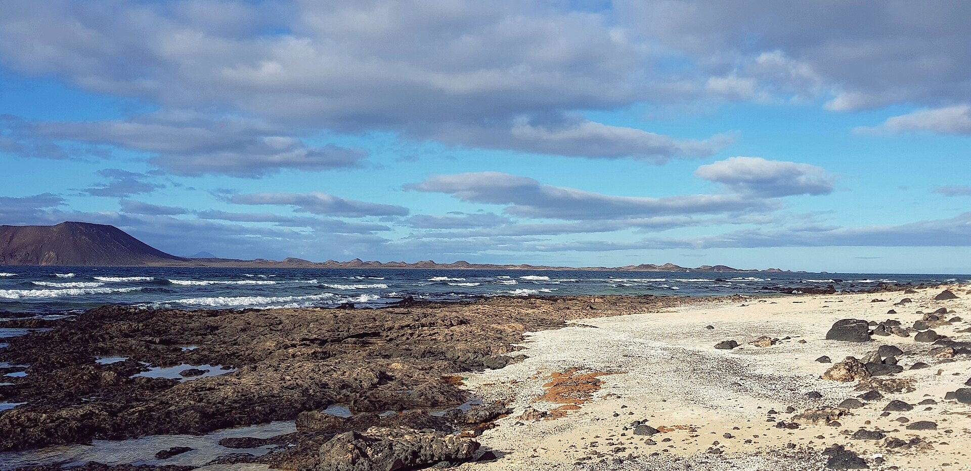Plaża w Corralejo na północy Fuerteventury: czarne skały wulkaniczne, jasny piasek i fale pod chmurami