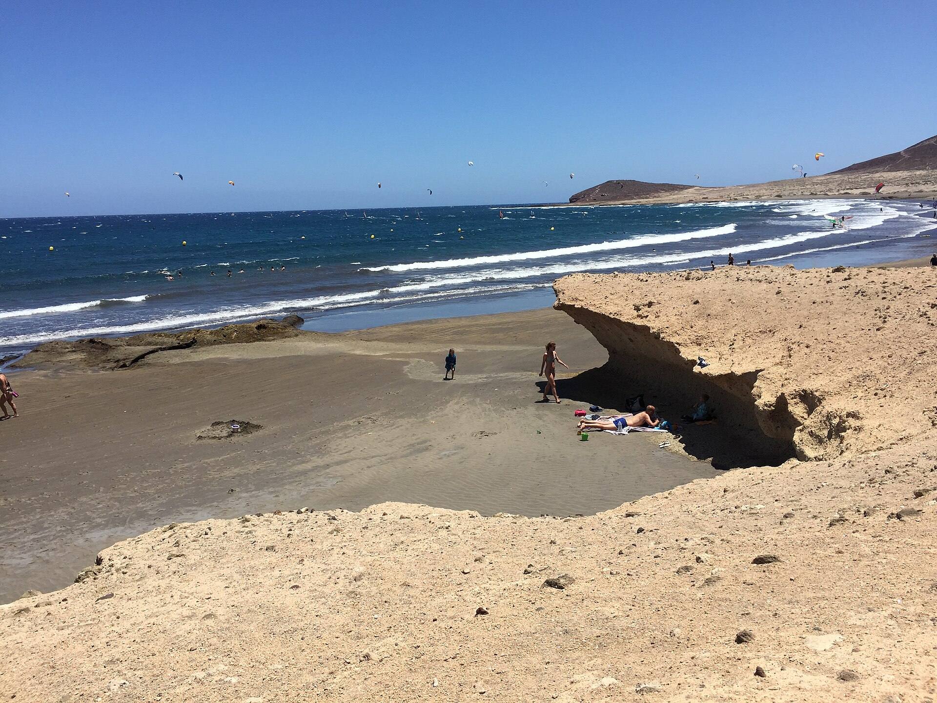 Plaża w El Médano na Teneryfie z jasnym skalnym nawisem na pierwszym planie, w tle góra Montaña Roja i latawce nad morzem