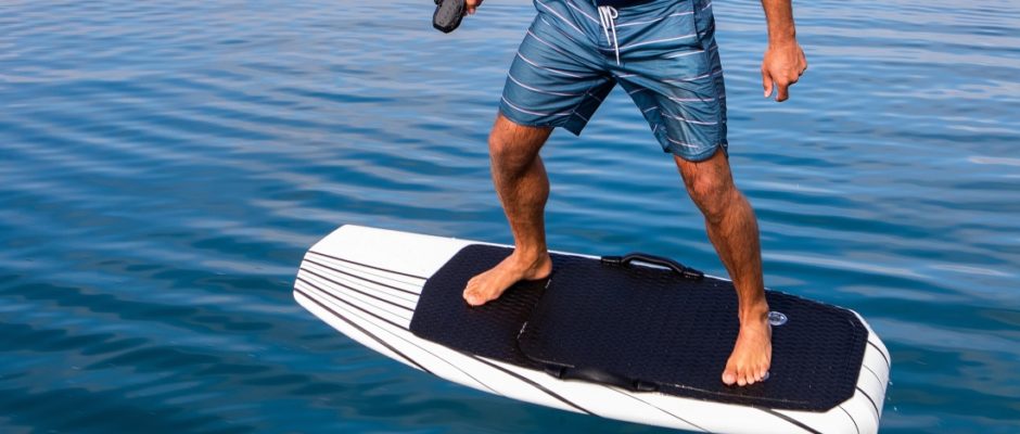 Barefoot rider in board shorts on a white eFoil board with a black deck pad, clear blue water all round; an illustration, not photographed in the Cayman Islands
