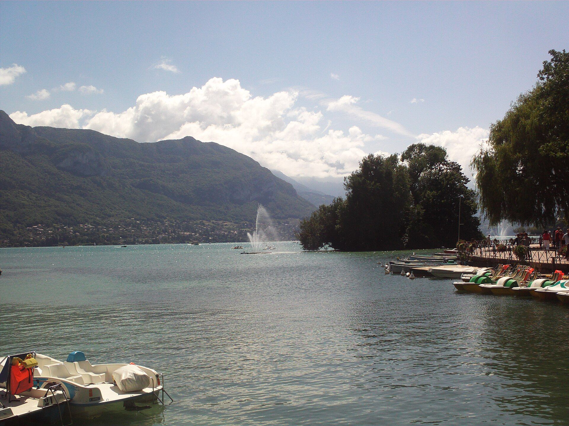 Lake Annecy with its fountain, pedal boats moored along a tree-lined quay on the right and wooded mountains on the far shore