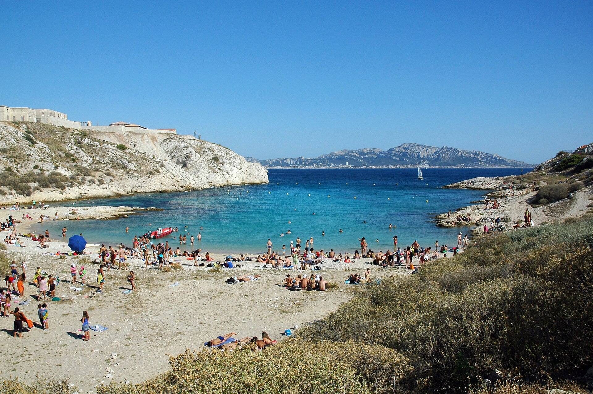 Swimmers at Saint-Estève beach, a small cove of turquoise water marked with buoys on Ratonneau island, with Marseille and its hills across the water