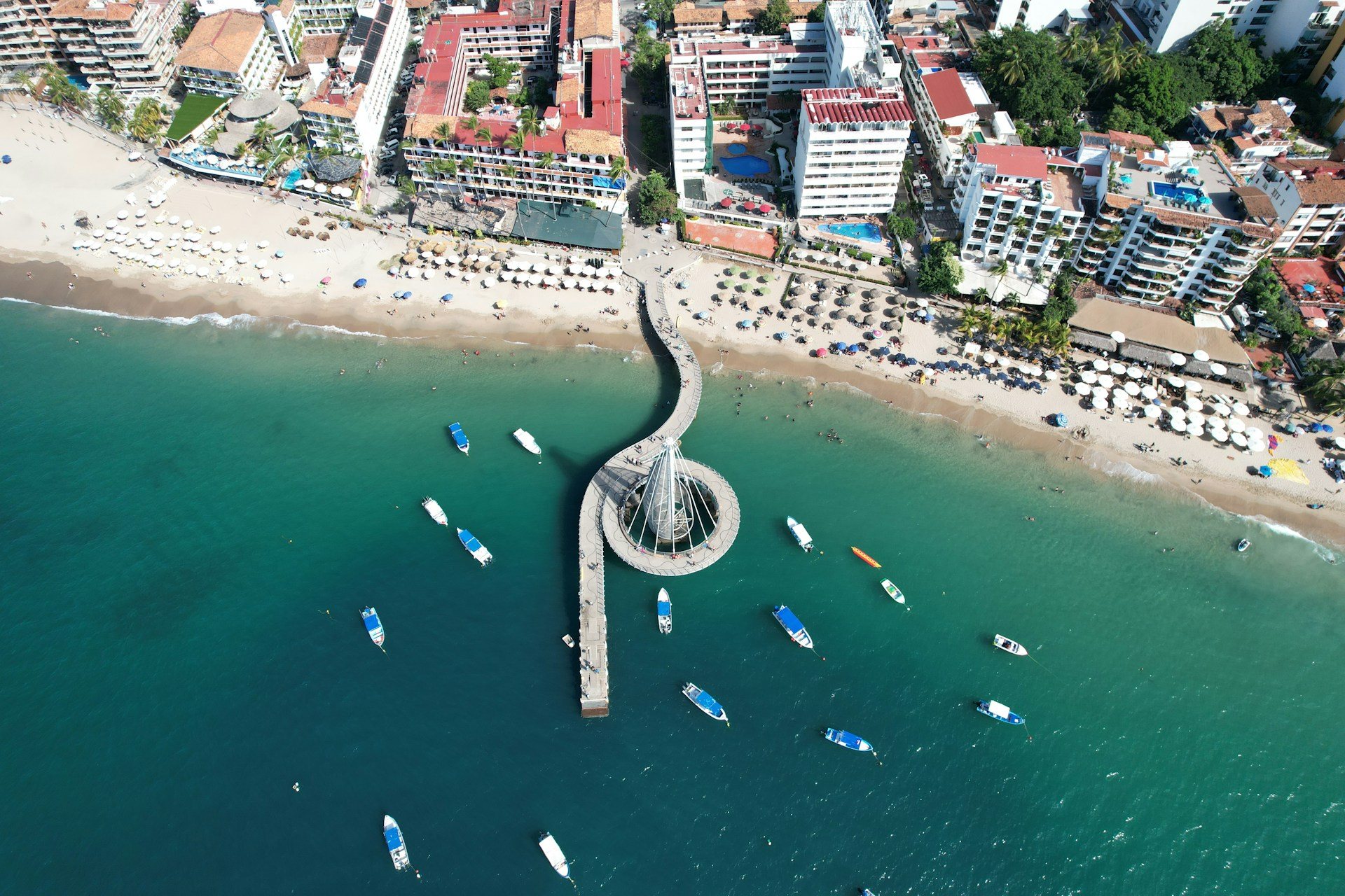 Aerial view of the Los Muertos pier in Puerto Vallarta, small boats moored around it and a beach of sunshades and hotels behind
